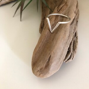K&ouml;nnte beinhalten: Ein silberner Ring mit einem V-f&ouml;rmigen Design. Der Ring liegt auf einem St&uuml;ck Treibholz.