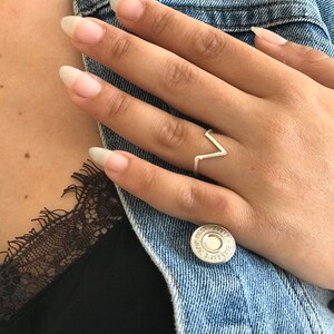 K&ouml;nnte beinhalten: Ein silberner Ring mit einem V-f&ouml;rmigen Design. Der Ring befindet sich an einem Finger.
