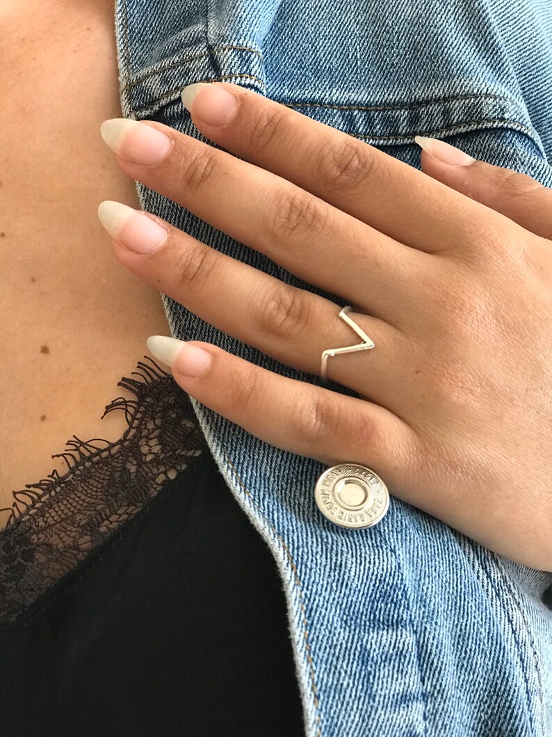 K&ouml;nnte beinhalten: Ein silberner Ring mit einem V-f&ouml;rmigen Design. Der Ring befindet sich an einem Finger.