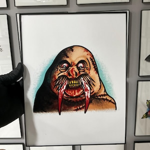 May include: A framed tattoo art print featuring a grotesque walrus with red tusks, yellow teeth, and a menacing expression. The walrus is depicted with a brown and tan body, surrounded by a light blue aura. The artwork is displayed among other framed tattoo designs.
