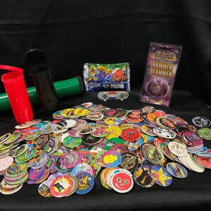 May include: A large collection of colorful plastic pogs with various designs, including cartoon characters, animals, and abstract patterns. There are also two small plastic containers, a package of pogs, and a purple and pink plastic toy with the text "Hammer Slammer" on the packaging.
