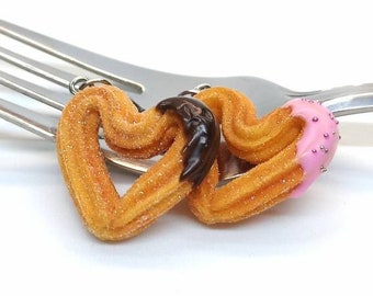 Könnte beinhalten: Zwei herzförmige Churros, einer mit Schokolade und der andere mit rosa Zuckerguss, werden auf einer silbernen Gabel aufgespießt.