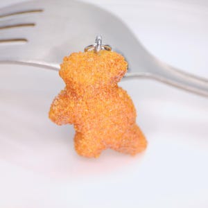 May include: A small, orange teddy bear charm with a textured surface, suspended from a silver ring. The charm is set against a white background with a silver fork.