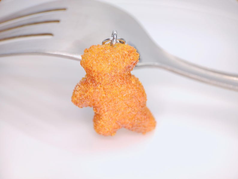 May include: A small, orange teddy bear charm with a textured surface, suspended from a silver ring. The charm is set against a white background with a silver fork.