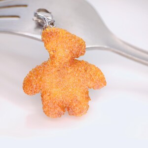 May include: A charm in the shape of a teddy bear made of orange material with a textured surface. The charm is attached to a silver clasp and is set against a silver fork.