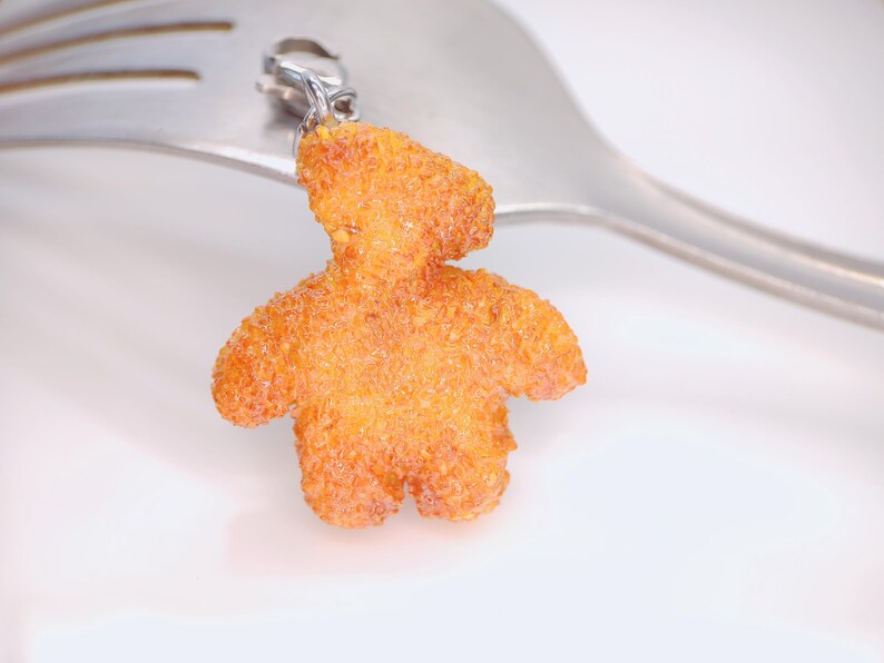 May include: A charm in the shape of a teddy bear made of orange material with a textured surface. The charm is attached to a silver clasp and is set against a silver fork.