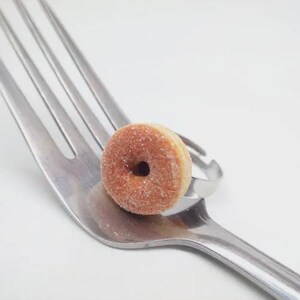 K&ouml;nnte beinhalten: Ein Miniatur-Donut-Ring mit Zuckerbeschichtung liegt auf einer silbernen Gabel. Der Ring ist hellbraun mit einem dunkelbraunen Loch in der Mitte. Die Gabel besteht aus gl&auml;nzendem Metall und hat vier Zinken.