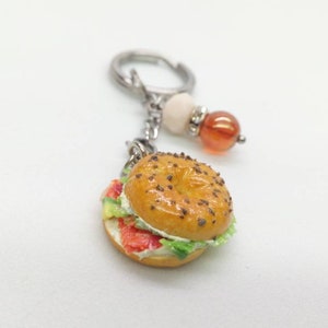 Pu&ograve; includere: Un portachiavi a forma di bagel in miniatura con un design realistico. Il bagel &egrave; ricoperto di semi di sesamo e farcito con ingredienti rossi, bianchi e verdi. Il portachiavi ha una chiusura color argento e perline decorative.