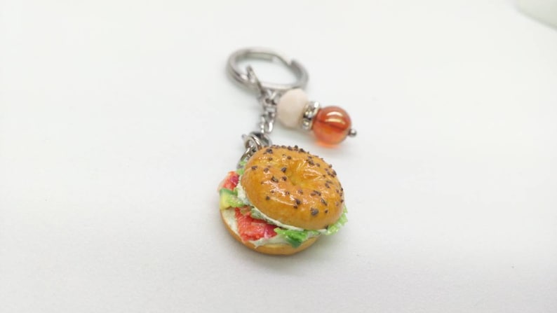Pu&ograve; includere: Un portachiavi a forma di bagel in miniatura con un design realistico. Il bagel &egrave; ricoperto di semi di sesamo e farcito con ingredienti rossi, bianchi e verdi. Il portachiavi ha una chiusura color argento e perline decorative.