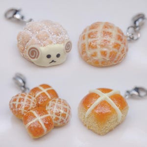 Puede incluir: Una colección de colgantes de comida en miniatura, que incluyen un pastel con forma de oveja, un pan redondo con diseño de enrejado y varios bollos de Pascua con cruces blancas. Cada colgante tiene un cierre plateado.