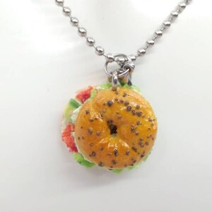 Pu&ograve; includere: Una collana con pendente con un ciondolo a forma di sandwich bagel in miniatura. Il bagel &egrave; marrone dorato con semi scuri e il sandwich include ripieni rossi, bianchi e verdi. La collana ha una catena a sfere color argento.