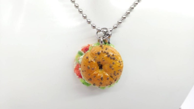Pu&ograve; includere: Una collana con pendente con un ciondolo a forma di sandwich bagel in miniatura. Il bagel &egrave; marrone dorato con semi scuri e il sandwich include ripieni rossi, bianchi e verdi. La collana ha una catena a sfere color argento.