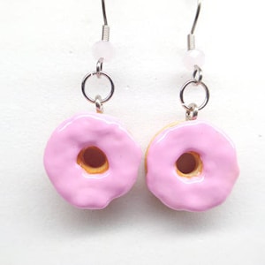 K&ouml;nnte beinhalten: Ein Paar Ohrh&auml;nger mit Donut-Anh&auml;ngern. Jeder Anh&auml;nger ist ein Miniatur-Donut mit rosa Glasur, brauner Mitte und gelbem Boden. Die Ohrringe haben silberfarbene Haken und eine kleine rosa Perle.