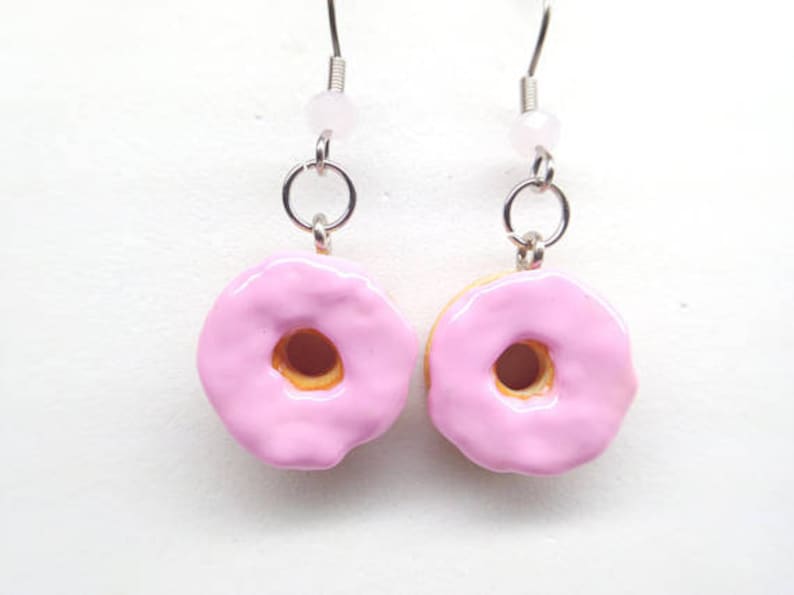 K&ouml;nnte beinhalten: Ein Paar Ohrh&auml;nger mit Donut-Anh&auml;ngern. Jeder Anh&auml;nger ist ein Miniatur-Donut mit rosa Glasur, brauner Mitte und gelbem Boden. Die Ohrringe haben silberfarbene Haken und eine kleine rosa Perle.