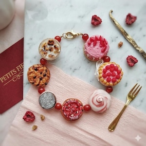 Könnte beinhalten: Ein einzigartiges Armband mit Miniatur-Dessert-Anhängern, darunter Kekse, Kuchen und Törtchen, die durch rote Perlen und einen goldenen Verschluss verbunden sind. Das Armband wird auf einer rosa Oberfläche mit einer goldenen Gabel und getrockneten Himbeeren präsentiert.