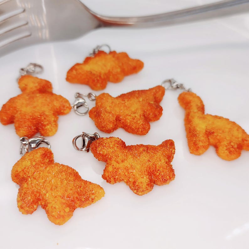 May include: Close-up of six orange, textured dinosaur-shaped charms with silver-colored clasps. The charms are arranged on a white surface. The charms are a bright orange color.