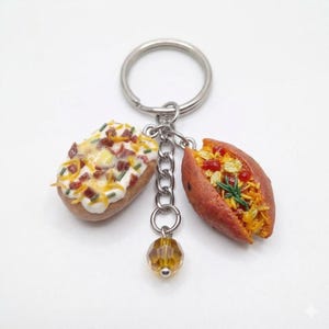 May include: A novelty keychain featuring miniature food charms. The charms include a baked potato with toppings and a sweet potato with filling. The keychain has a silver-colored ring, chain, and a decorative amber-colored bead.