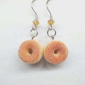 K&ouml;nnte beinhalten: Ein Paar ausgefallene Ohrringe mit Miniatur-Donut-Anh&auml;ngern. Jeder Ohrring hat einen silberfarbenen Haken, eine kleine gelbe Perle und einen Ring, der mit einem Donut-f&ouml;rmigen Anh&auml;nger verbunden ist. Die Donuts sind hellbraun mit einer zuckerartigen Beschichtung.