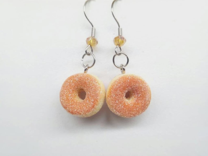 K&ouml;nnte beinhalten: Ein Paar ausgefallene Ohrringe mit Miniatur-Donut-Anh&auml;ngern. Jeder Ohrring hat einen silberfarbenen Haken, eine kleine gelbe Perle und einen Ring, der mit einem Donut-f&ouml;rmigen Anh&auml;nger verbunden ist. Die Donuts sind hellbraun mit einer zuckerartigen Beschichtung.