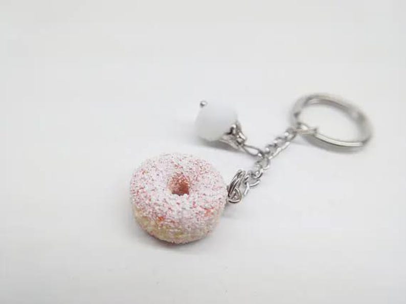 K&ouml;nnte beinhalten: Ein Schl&uuml;sselanh&auml;nger in Donut-Form mit rosa Zuckerguss und wei&szlig;en Streuseln. Der Schl&uuml;sselanh&auml;nger hat eine silberne Kette, eine wei&szlig;e Perle und einen silbernen Schl&uuml;sselring. Der Donut ist eine Miniatur.