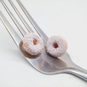 K&ouml;nnte beinhalten: Zwei Mini-Donuts, best&auml;ubt mit Puderzucker, liegen auf einer silbernen Gabel vor wei&szlig;em Hintergrund. Die Donuts sind hellbraun mit einem Loch in der Mitte. Die Zinken der Gabel sind sichtbar und bilden eine diagonale Linie.