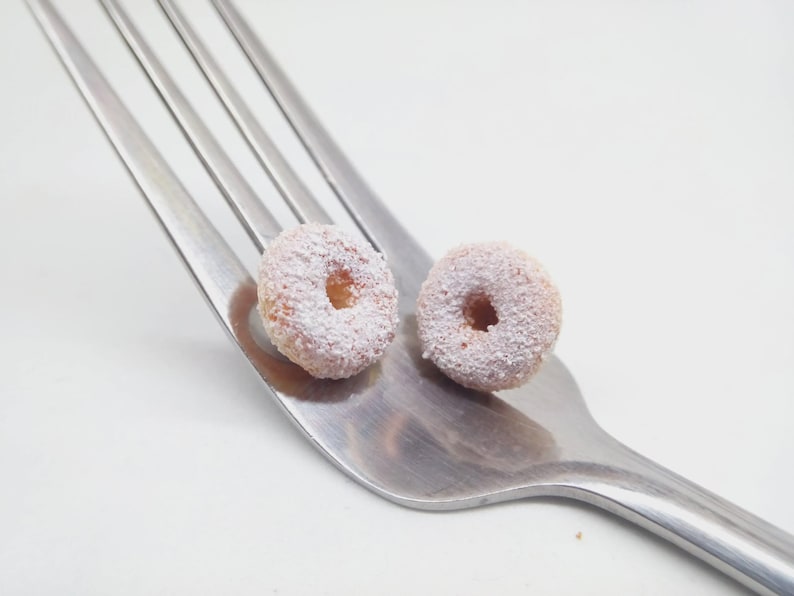 K&ouml;nnte beinhalten: Zwei Mini-Donuts, best&auml;ubt mit Puderzucker, liegen auf einer silbernen Gabel vor wei&szlig;em Hintergrund. Die Donuts sind hellbraun mit einem Loch in der Mitte. Die Zinken der Gabel sind sichtbar und bilden eine diagonale Linie.