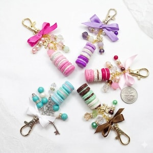 May include: A collection of keyrings with miniature macarons in shades of pink, purple, blue, and brown. Each keyring has a gold-tone clasp, decorative beads, and a ribbon bow. Some keyrings include a small key charm.