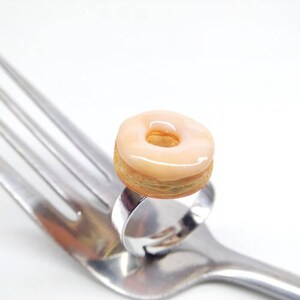 K&ouml;nnte beinhalten: Ein Miniatur-Donut-Ring mit hellrosa Glasur liegt auf einer silbernen Gabel. Der Donut hat einen goldbraunen Geb&auml;ckboden und ein Loch in der Mitte. Der Hintergrund ist schlicht wei&szlig;.