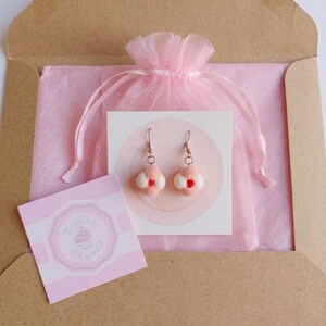 Pu&ograve; includere: Un paio di orecchini a forma di fiore rosa e bianco con centri rossi sono esposti su una carta bianca all'interno di un sacchetto di organza rosa. Gli orecchini sono confezionati in una busta marrone con carta velina rosa. &Egrave; inclusa anche una piccola carta rosa con un logo di cupcake.