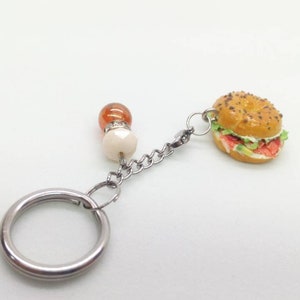 Pu&ograve; includere: Un portachiavi a forma di hamburger in miniatura con catena e anello argentati. L'hamburger ha un panino al sesamo, lattuga, pomodoro e un hamburger. La catena &egrave; collegata a perline arancioni e bianche.
