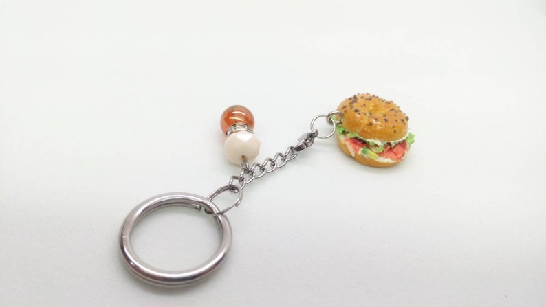 Pu&ograve; includere: Un portachiavi a forma di hamburger in miniatura con catena e anello argentati. L'hamburger ha un panino al sesamo, lattuga, pomodoro e un hamburger. La catena &egrave; collegata a perline arancioni e bianche.