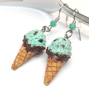 Puede incluir: Un par de pendientes de cono de helado. Cada pendiente presenta una bola de helado verde menta con chispas de chocolate sobre un cono de gofre marrón. El helado está rociado con chocolate. Los pendientes cuelgan de ganchos plateados con cuentas verdes.