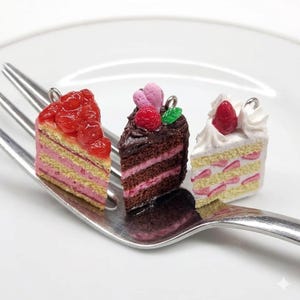 以下が含まれることがあります： フォークに乗った3つのミニチュアケーキスライス。スライスは、イチゴ、チョコレート、ストロベリーショートケーキのデザインが特徴です。各ケーキには、吊り下げ用のシルバーのループが付いています。ケーキは白いお皿の上にあります。