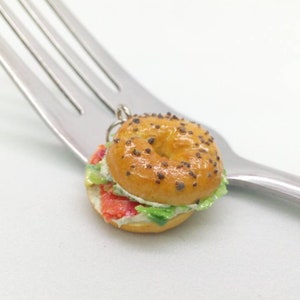 Pu&ograve; includere: Un ciondolo a forma di bagel in miniatura con un design realistico, con un esterno marrone dorato cosparso di semi scuri. Il bagel &egrave; tagliato e farcito con ingredienti rossi, bianchi e verdi, appoggiato su una forchetta d'argento. Un accessorio a tema cibo.