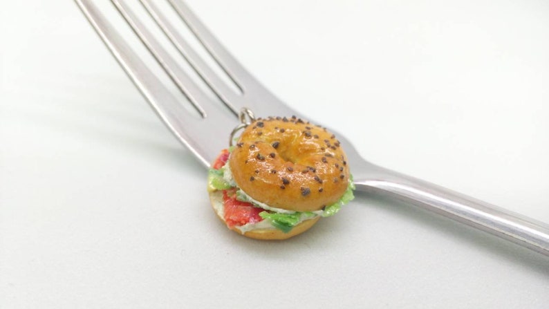 Pu&ograve; includere: Un ciondolo a forma di bagel in miniatura con un design realistico, con un esterno marrone dorato cosparso di semi scuri. Il bagel &egrave; tagliato e farcito con ingredienti rossi, bianchi e verdi, appoggiato su una forchetta d'argento. Un accessorio a tema cibo.