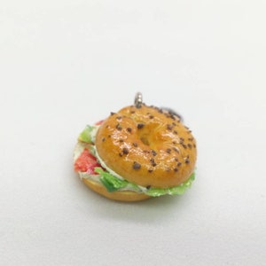 Pu&ograve; includere: Un ciondolo a forma di bagel in miniatura con una parte superiore lucida marrone dorato cosparsa di semi scuri. Il bagel &egrave; farcito con strati di ingredienti rossi, bianchi e verdi, che assomigliano a un panino. Il ciondolo &egrave; su sfondo bianco.