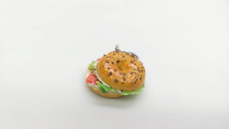 Pu&ograve; includere: Un ciondolo a forma di bagel in miniatura con una parte superiore lucida marrone dorato cosparsa di semi scuri. Il bagel &egrave; farcito con strati di ingredienti rossi, bianchi e verdi, che assomigliano a un panino. Il ciondolo &egrave; su sfondo bianco.