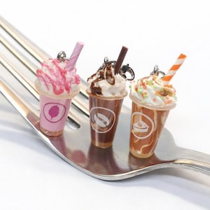 May include: Three miniature iced coffee charms with whipped cream and straws sit on a silver fork. The charms are in pink, brown, and tan, each with a different logo. The pink charm has a pink and white striped straw.