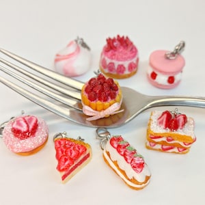 Könnte beinhalten: Eine Sammlung miniaturer Dessert-Anhänger, darunter ein rosa Macaron, ein Erdbeerkuchen und ein Stück Erdbeertarte. Die Anhänger sind auf einer silbernen Gabel vor weißem Hintergrund angeordnet. Jeder Anhänger hat einen silbernen Verschluss.
