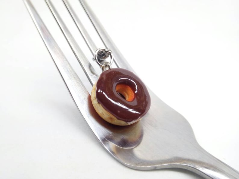 K&ouml;nnte beinhalten: Ein Miniatur-Donut-Anh&auml;nger mit Schokoladenglasur, der auf einer Silber-Gabel liegt. Der Donut hat eine hellbraune Basis und einen dunkelbraunen, gl&auml;nzenden Belag. Der Anh&auml;nger ist an einer kleinen Silberschlaufe befestigt.