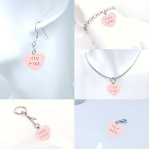 May include: A collection of jewelry items featuring pink heart-shaped charms with the text "ITEM HERE". The items include earrings, a bracelet, a necklace, a keychain, and a charm. The charms are attached to silver-colored metal chains and findings.