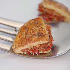 Könnte beinhalten: Nahaufnahme eines Mini-Sandwiches auf einer Silberforke. Das Sandwich hat eine goldbraune, strukturierte Brotoberseite und ist mit roten und weißen Gemüsen und Fleisch gefüllt. Ein weiteres Sandwich ist im Hintergrund leicht unscharf.