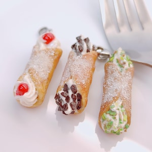 Puede incluir: Tres miniaturas de pasteles cannoli con diferentes coberturas. Uno tiene una cereza, otro chispas de chocolate y el tercero chispas verdes. Todos están espolvoreados con azúcar glas y sobre una superficie blanca.