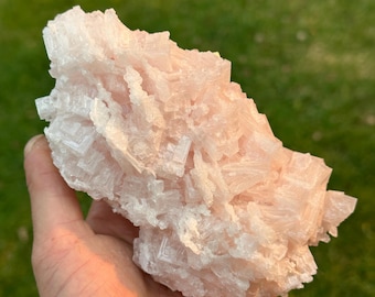 May include: A large, light pink crystal cluster with a rough, textured surface. The crystal is held in a hand against a blurred green background, showcasing its natural, geometric formations. The crystal's color ranges from pale to a deeper pink.