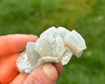 May include: A cluster of pale blue-green mineral crystals, held in a hand against a blurred green background. The crystals have a layered, geometric structure, with a slightly translucent appearance. The overall effect is delicate and natural.
