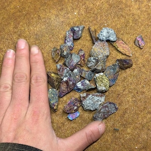 May include: A collection of iridescent pyrite stones, displaying a spectrum of colors including gold, purple, and blue. The rough-cut stones vary in size and shape, with a metallic sheen. The stones are on a brown surface.