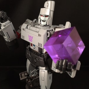 May include: A gray and black action figure of Megatron holding a large, purple, geometric cube. The figure has a silver face with red eyes and a purple emblem on the chest. The background is black.