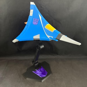 Op de afbeelding: Een blauw en wit speelgoedvliegtuig met een paars embleem op de vleugel. Het vliegtuig is gemonteerd op een zwarte standaard met een paars embleem op de basis. Het vliegtuig heeft gele en zilveren accenten.