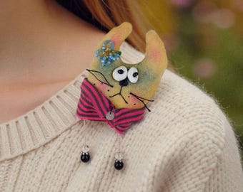 May include: A whimsical cat brooch made of fabric, featuring a yellow body, pink and black striped bow tie, and large, expressive eyes. The cat has a small blue flower on its head and is pinned to a cream-colored sweater. Two small black bead earrings hang below.