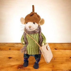 May include: A felt mouse wearing a brown hat and green sweater, carrying a brown bag. The mouse is standing on a wooden surface.
